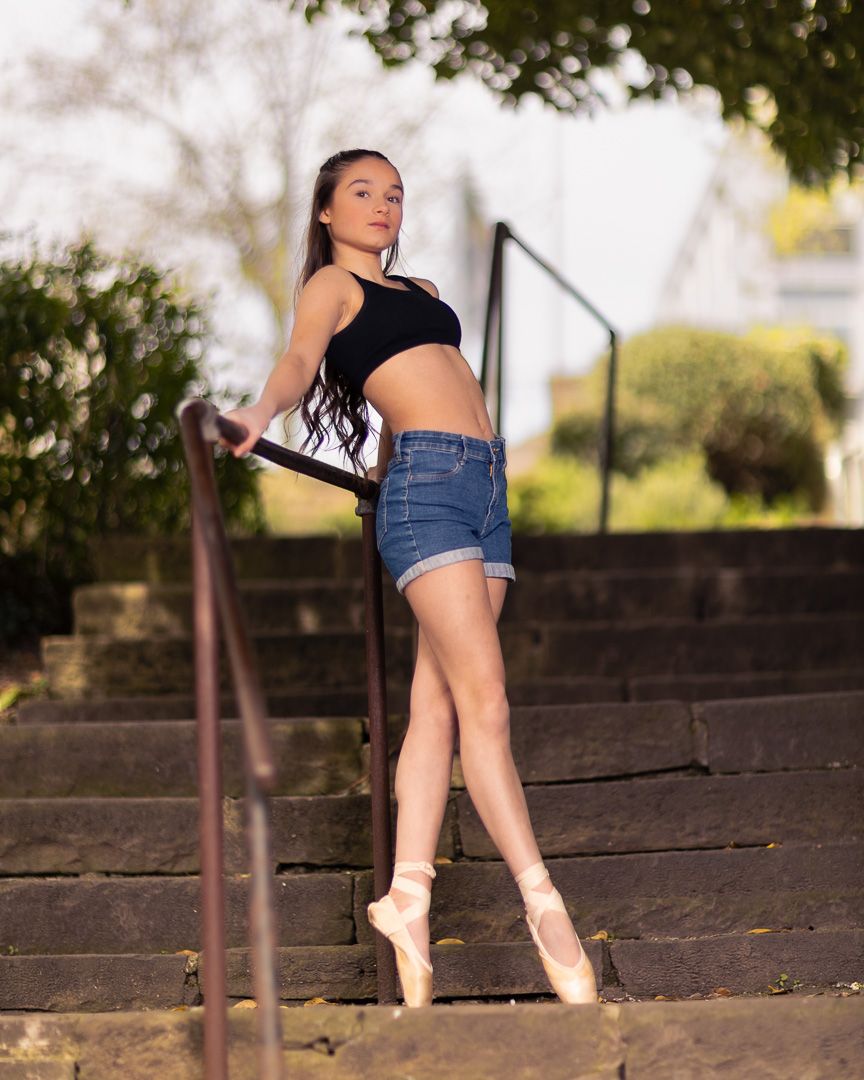 A young ballerina poses gracefully on outdoor steps in denim shorts and pointe shoes, holding a railing.