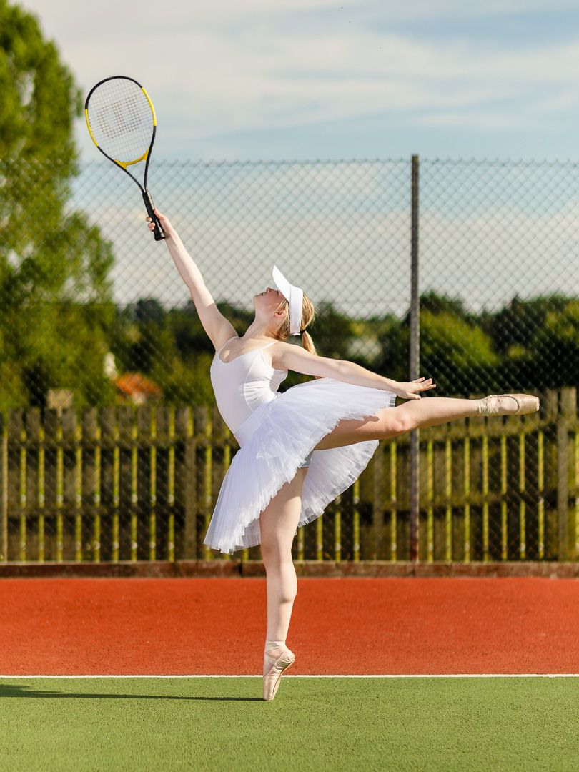 Ballerina in white tutu performs arabesque while holding tennis racket on outdoor court.