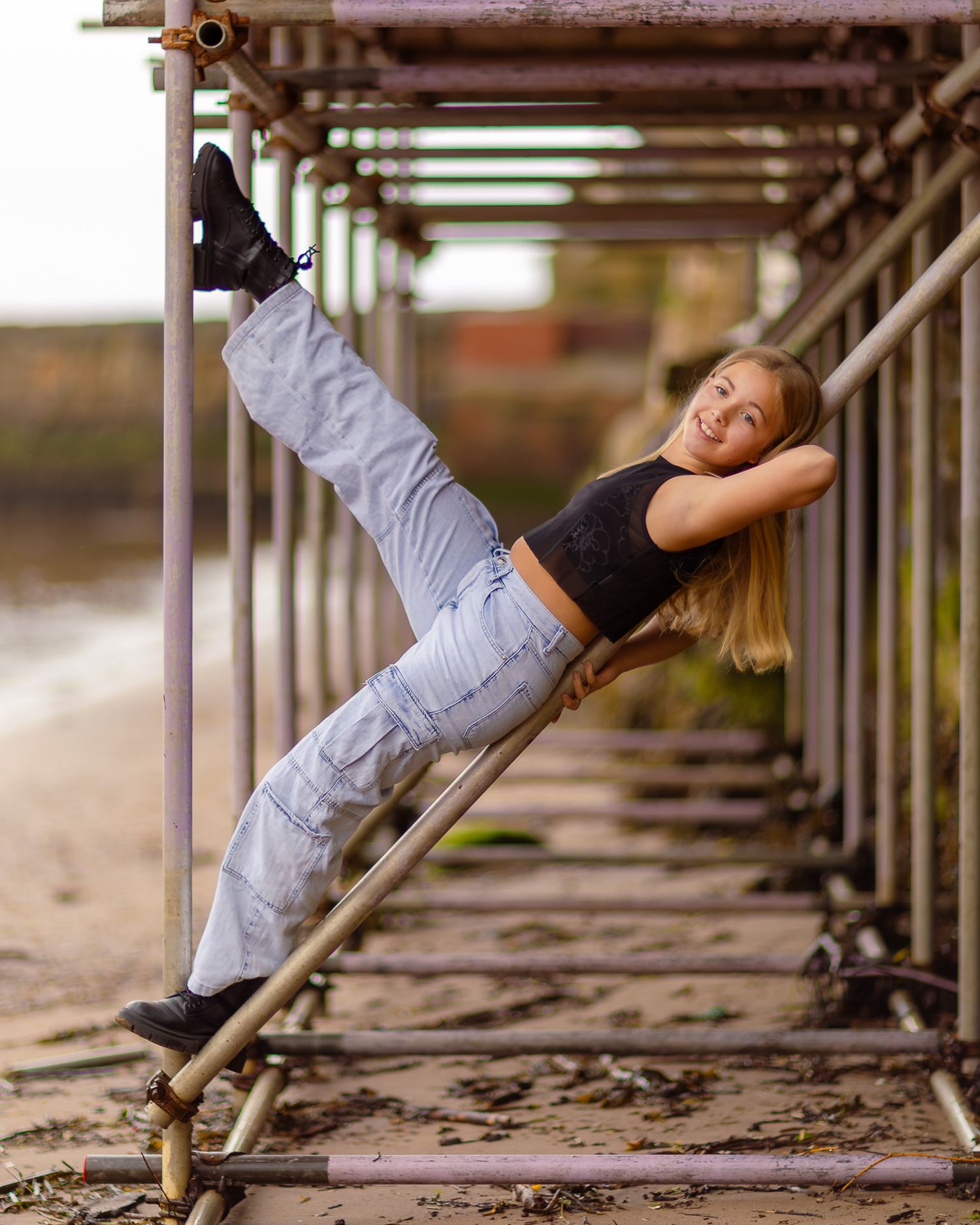 A flexible young girl poses dynamically on metal scaffolding near a beach, wearing a crop top and gray cargo pants.