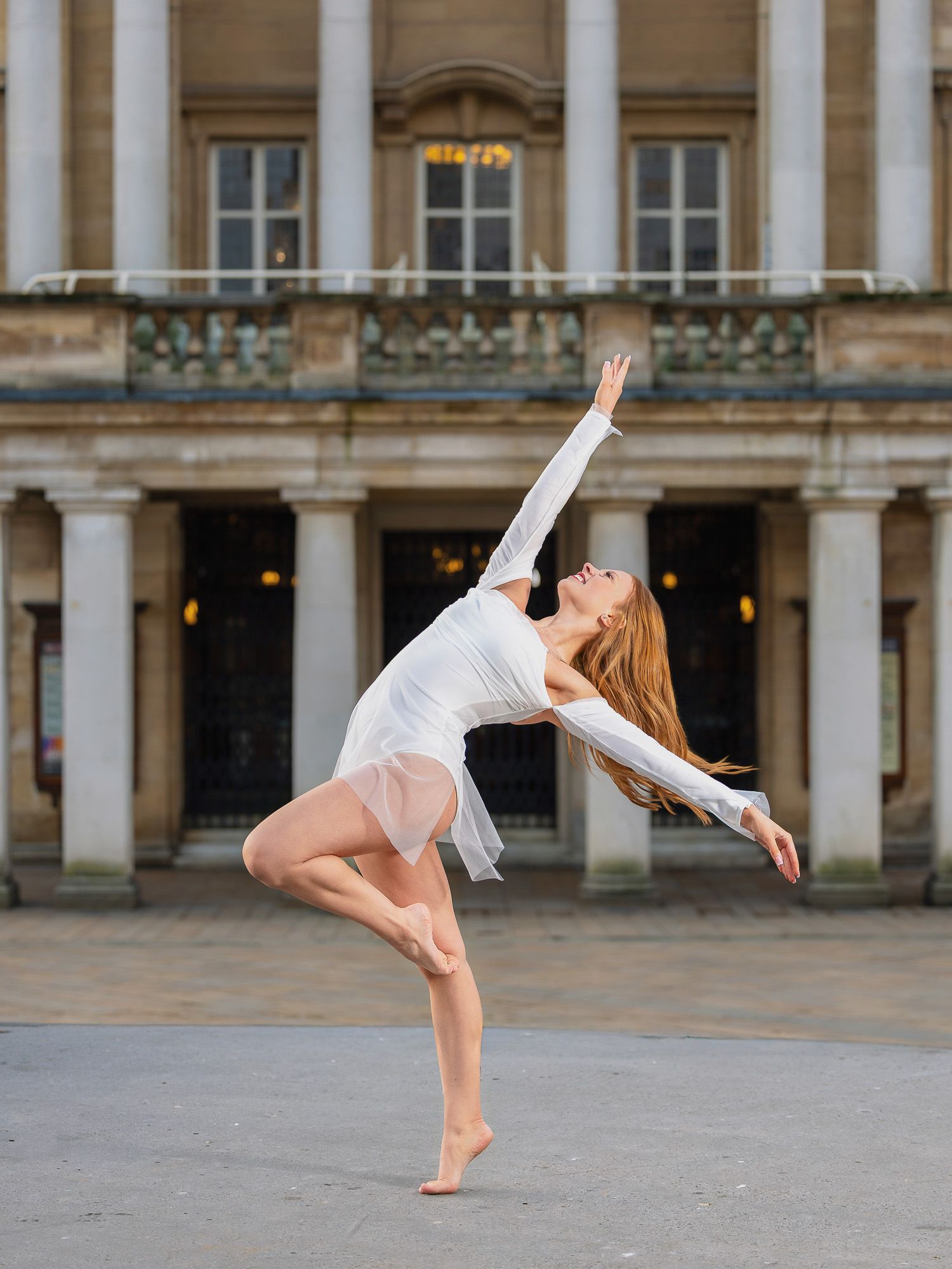 A ballet dancer in a white dress performs an elegant arabesque pose outdoors in front of a classical stone building.