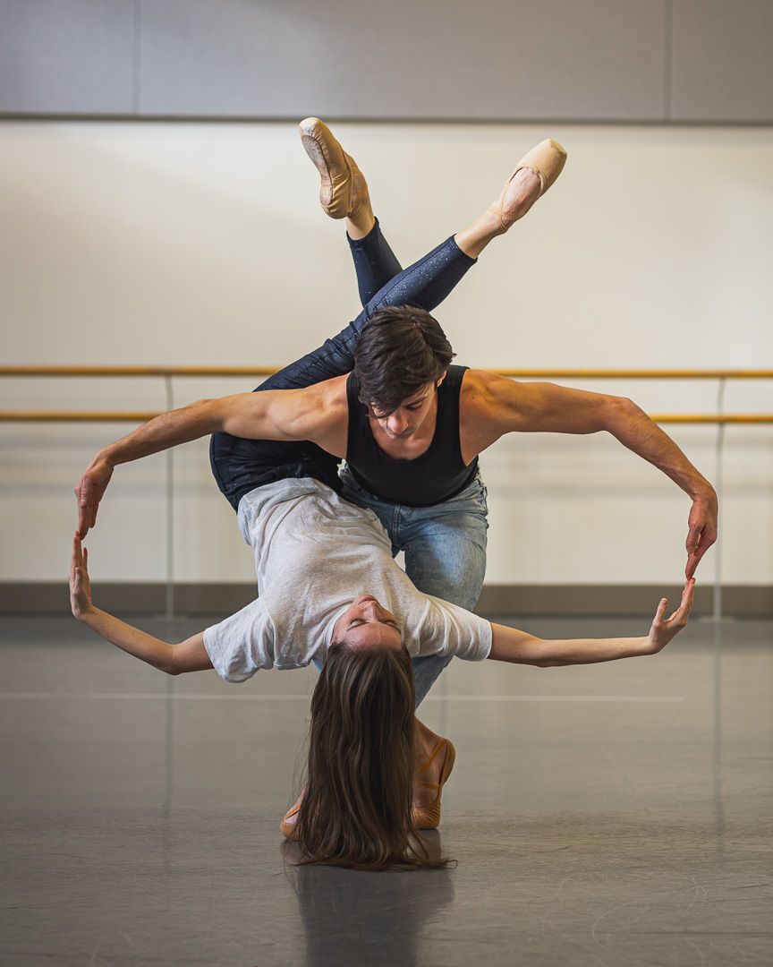 Two ballet dancers perform an acrobatic lift in a studio, with the female dancer inverted and both forming a circular shape.