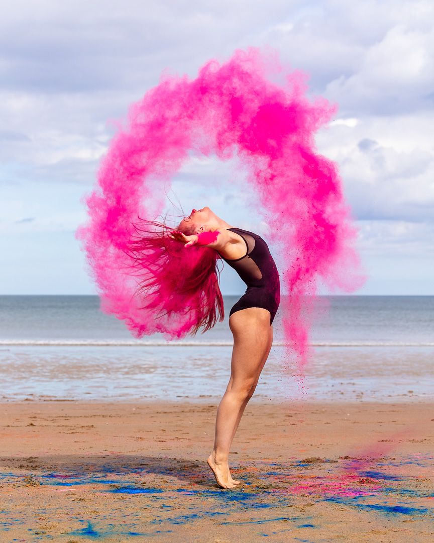 A dancer on a beach creates a stunning pink smoke arc while flipping her hair, wearing a black leotard.