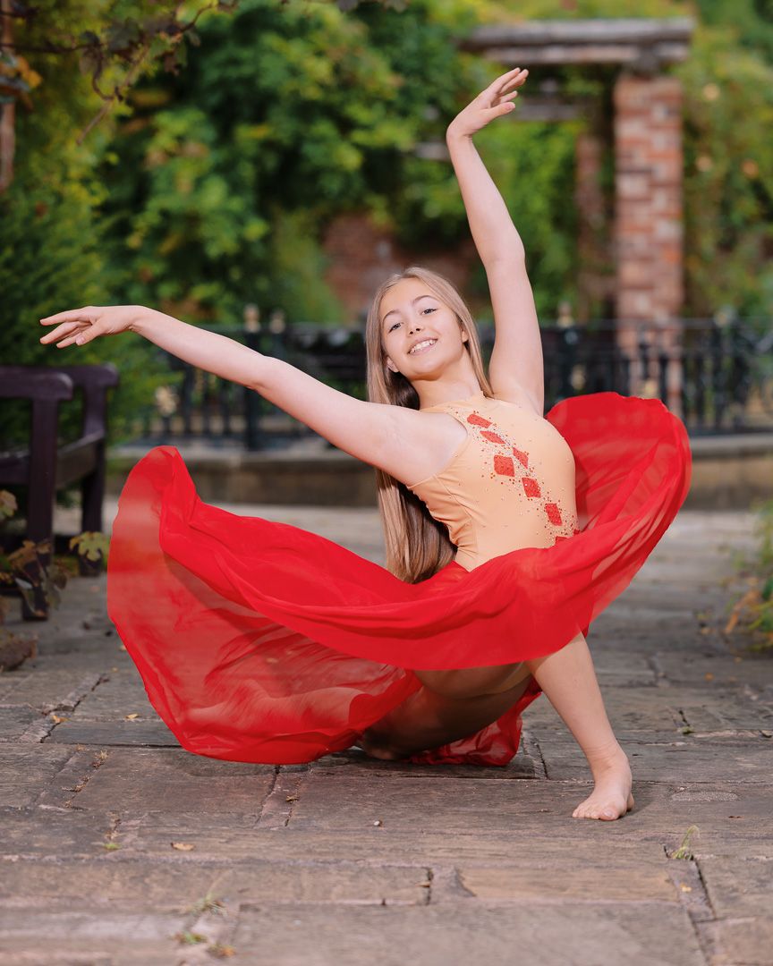 Young female dancer in red flowing skirt and orange leotard performs graceful arabesque on stone patio outdoors.