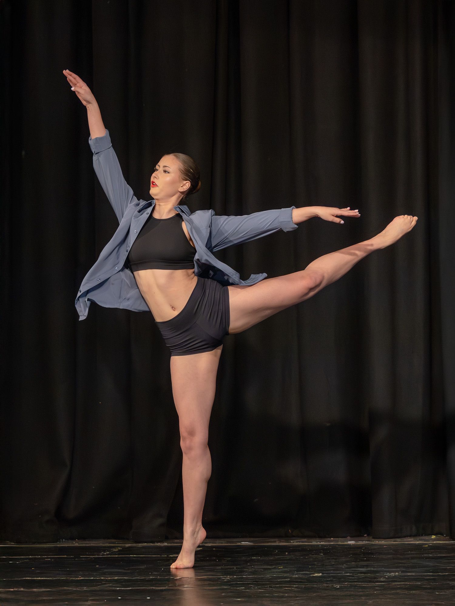 A dancer performs an arabesque on stage wearing a black crop top, shorts, and blue jacket under dramatic lighting.