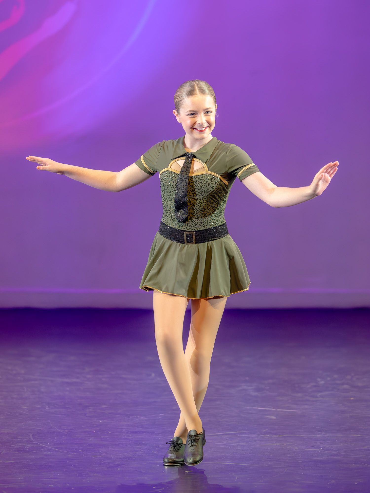 Young dancer in olive green costume performs tap dance on stage with purple backdrop.
