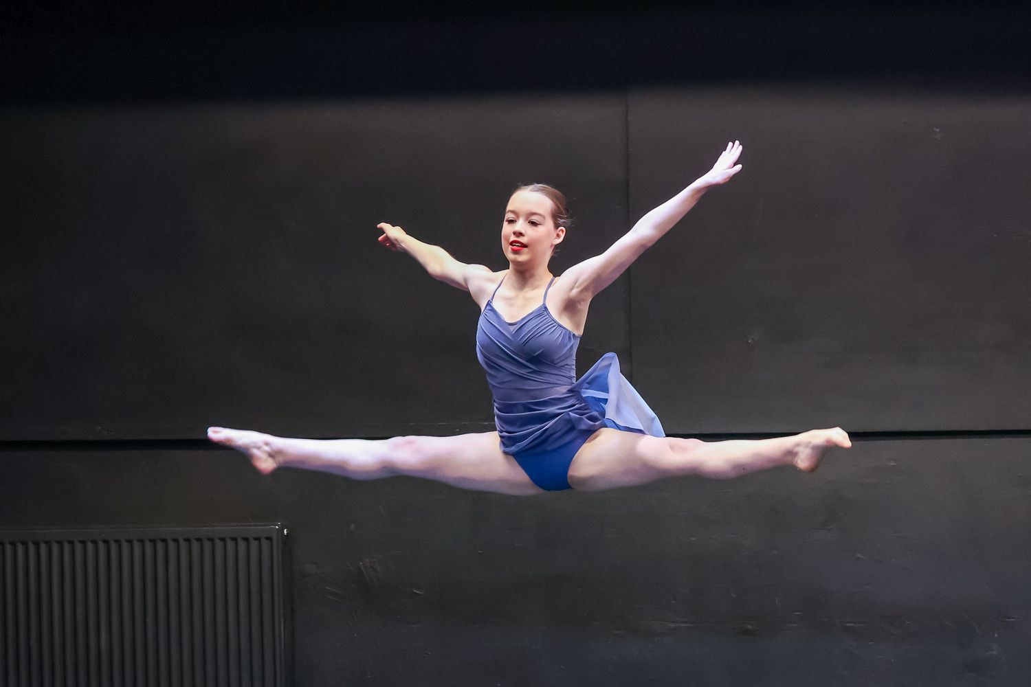 A female dancer leaps into a perfect split mid-air wearing a blue leotard during a ballet performance.