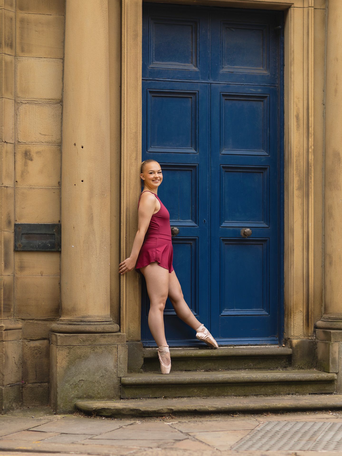 Graceful ballerina in burgundy leotard poses en pointe beside a bold blue door on stone steps.