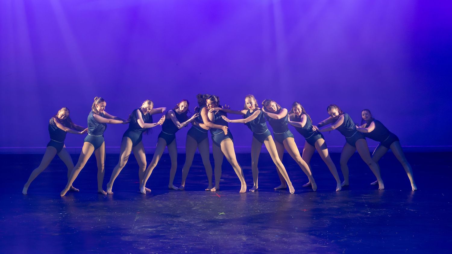 Group of dancers performing a synchronized routine on stage under purple lighting in dark costumes.