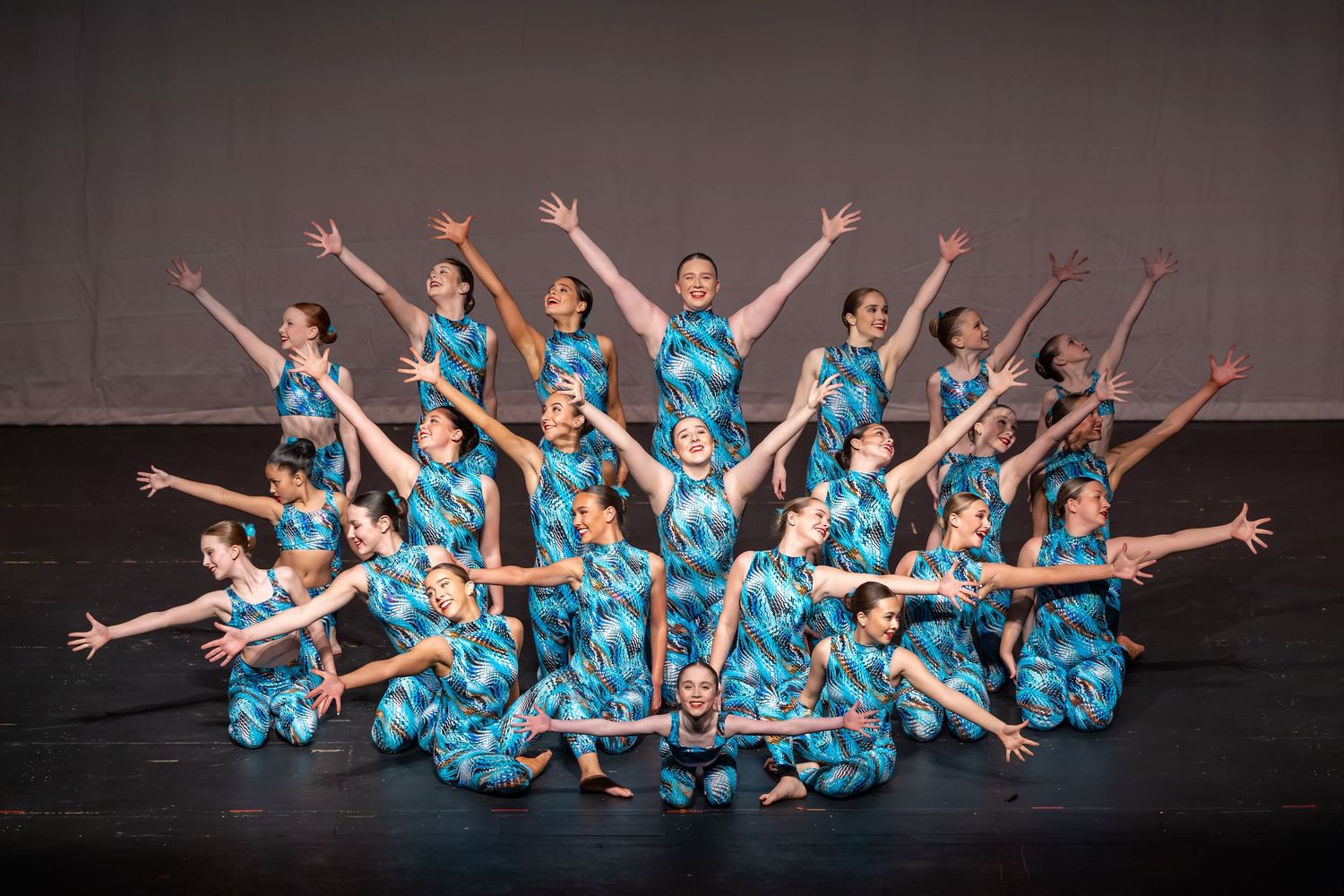 Large dance group performing on stage in matching teal blue patterned jumpsuits with arms raised in formation.