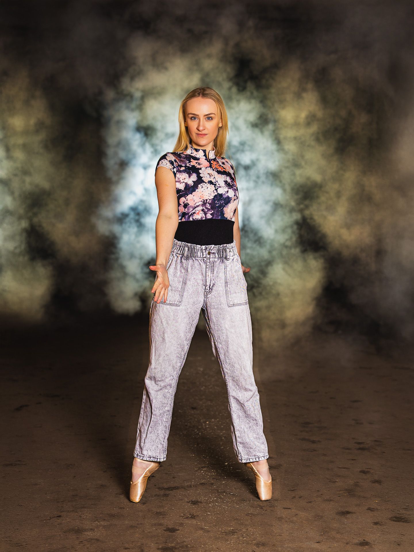 Blonde dancer in floral top, acid wash jeans, and ballet pointe shoes poses in a dramatic smoky studio.