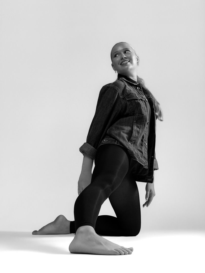 Bald woman in denim jacket and black leggings kneels gracefully in a black and white studio photo.