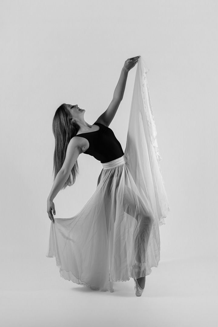 Black and white photo of a ballet dancer in a flowing white skirt and black top, gracefully posing on pointe.