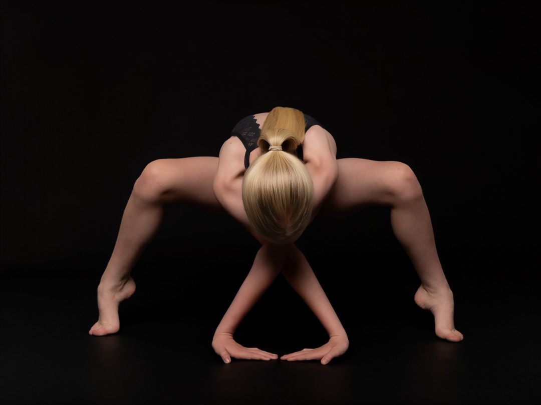 Blonde dancer in a black leotard performs a deep forward bend with hands and feet on a dark studio floor.