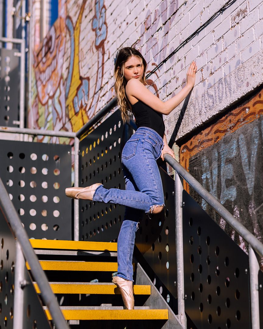 A young woman in jeans and a black top poses gracefully on yellow metal stairs near a graffiti wall.