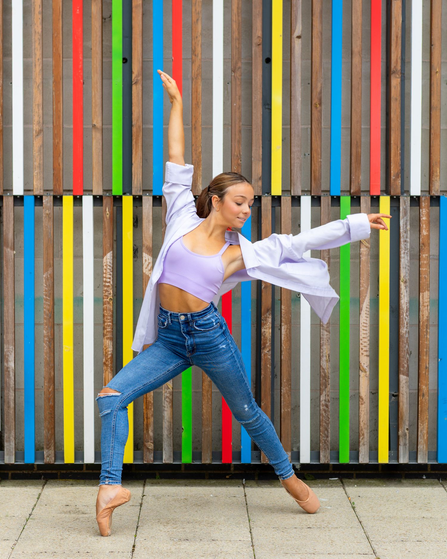 Dancer in blue jeans and white crop top strikes graceful pose against colorful striped wall background.