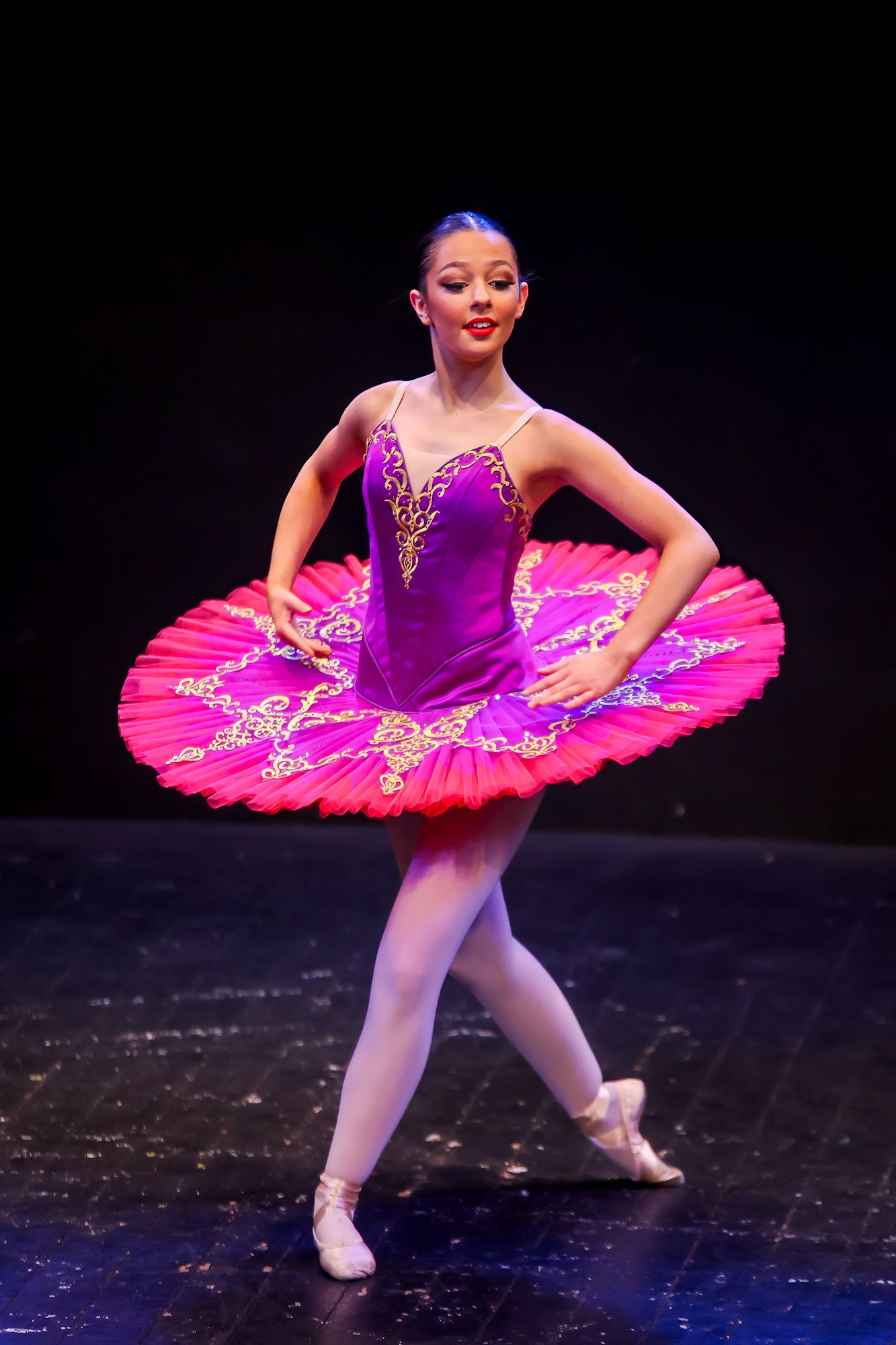 Young ballerina in pink and purple tutu performs on stage in classic ballet pose wearing pointe shoes.