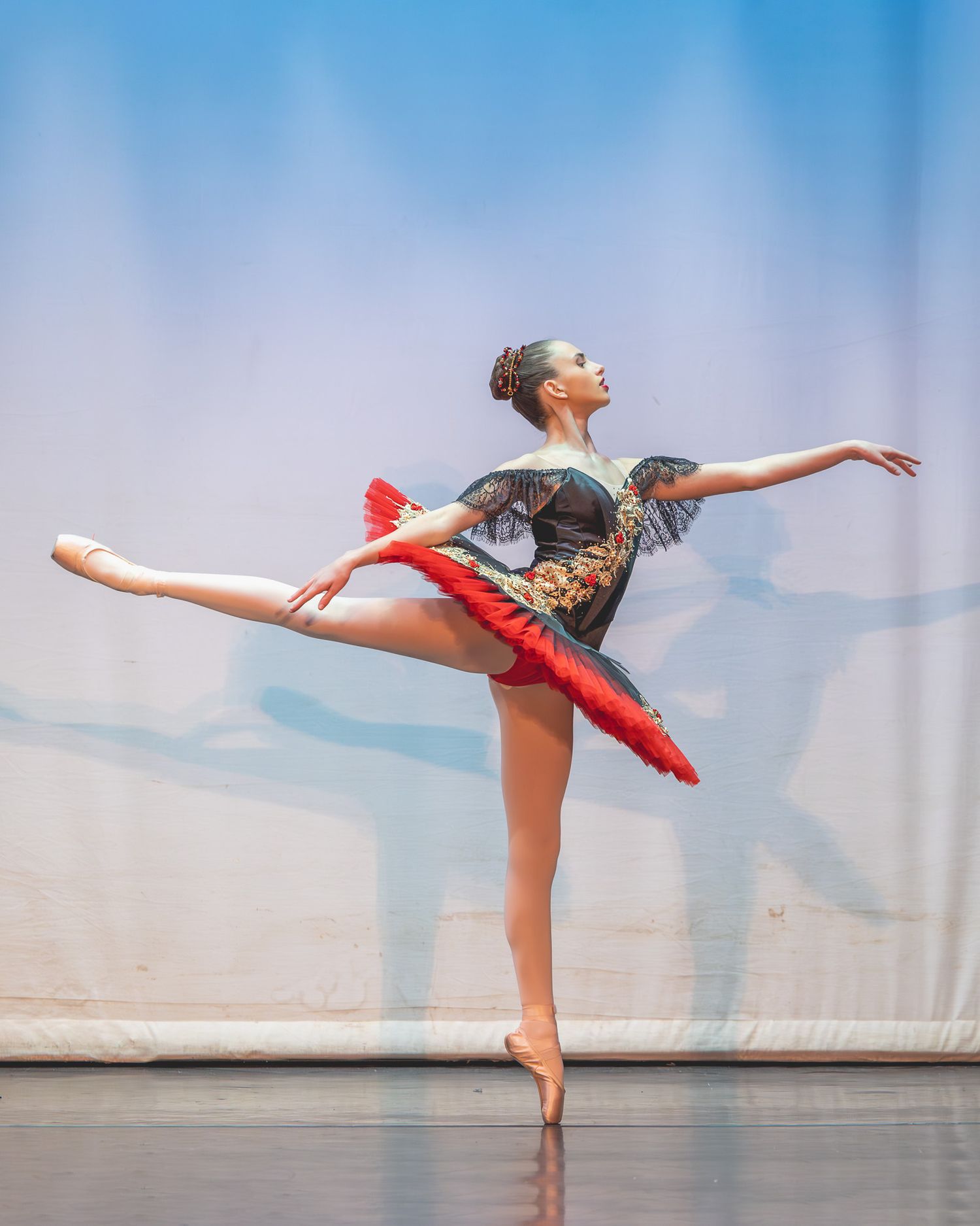Young ballerina performs arabesque on stage in red and black costume under blue spotlight.