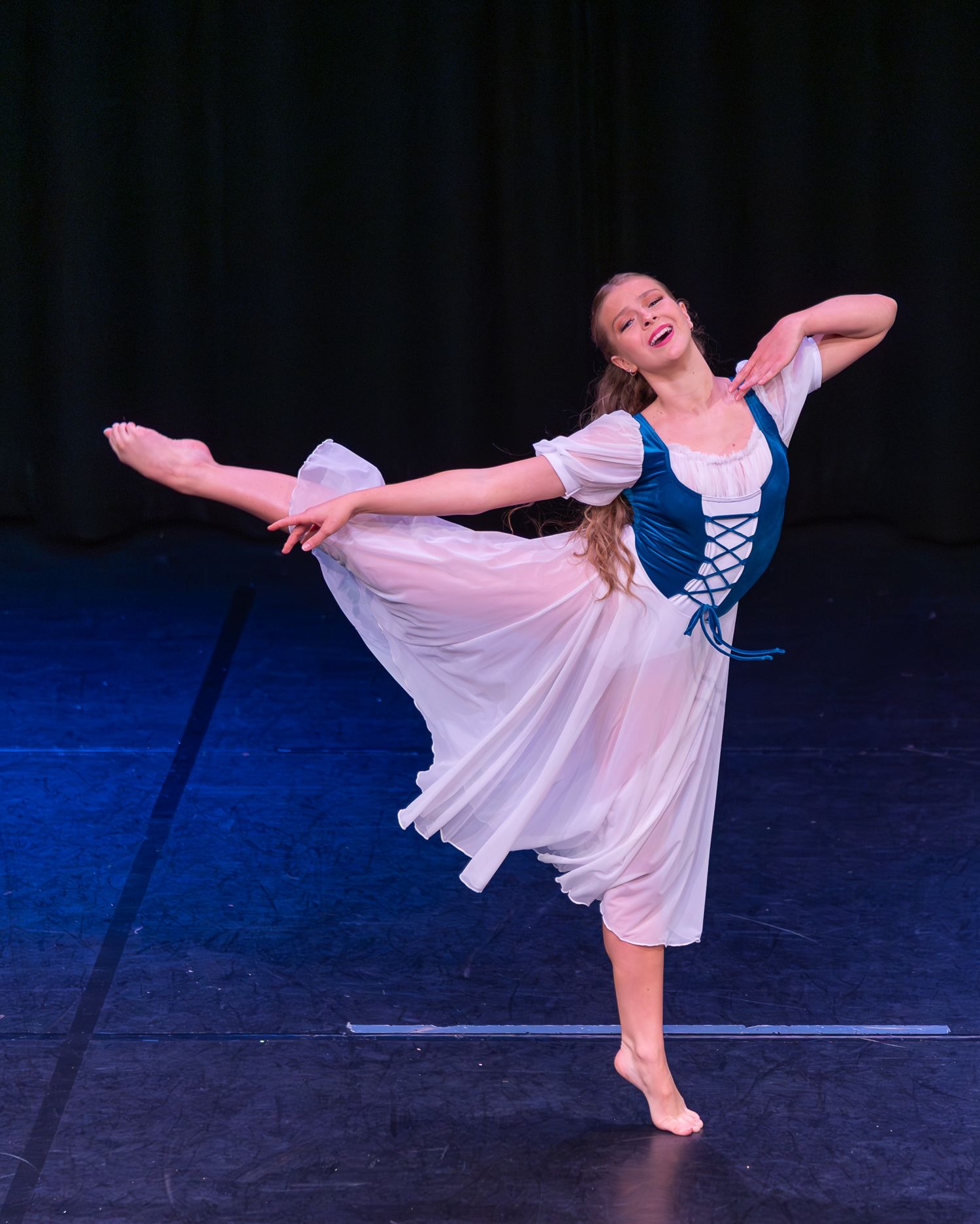Graceful ballet dancer in white and teal costume performs an arabesque on stage under blue lighting.