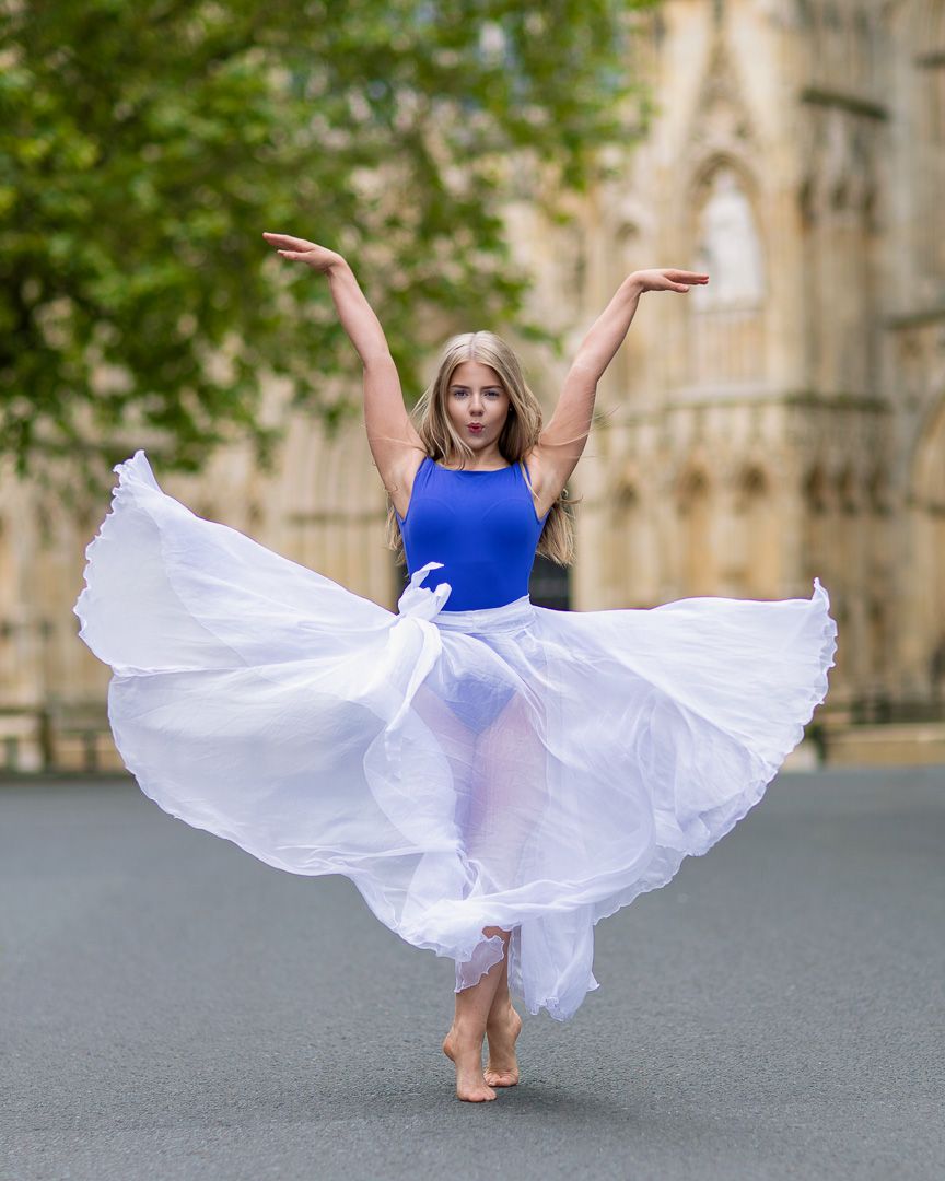Dancer in blue leotard twirls in flowing white dress against stone architecture.