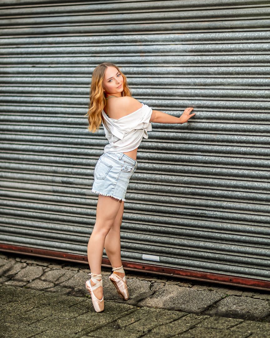 Dancer in white top and denim skirt poses en pointe against industrial metal backdrop.