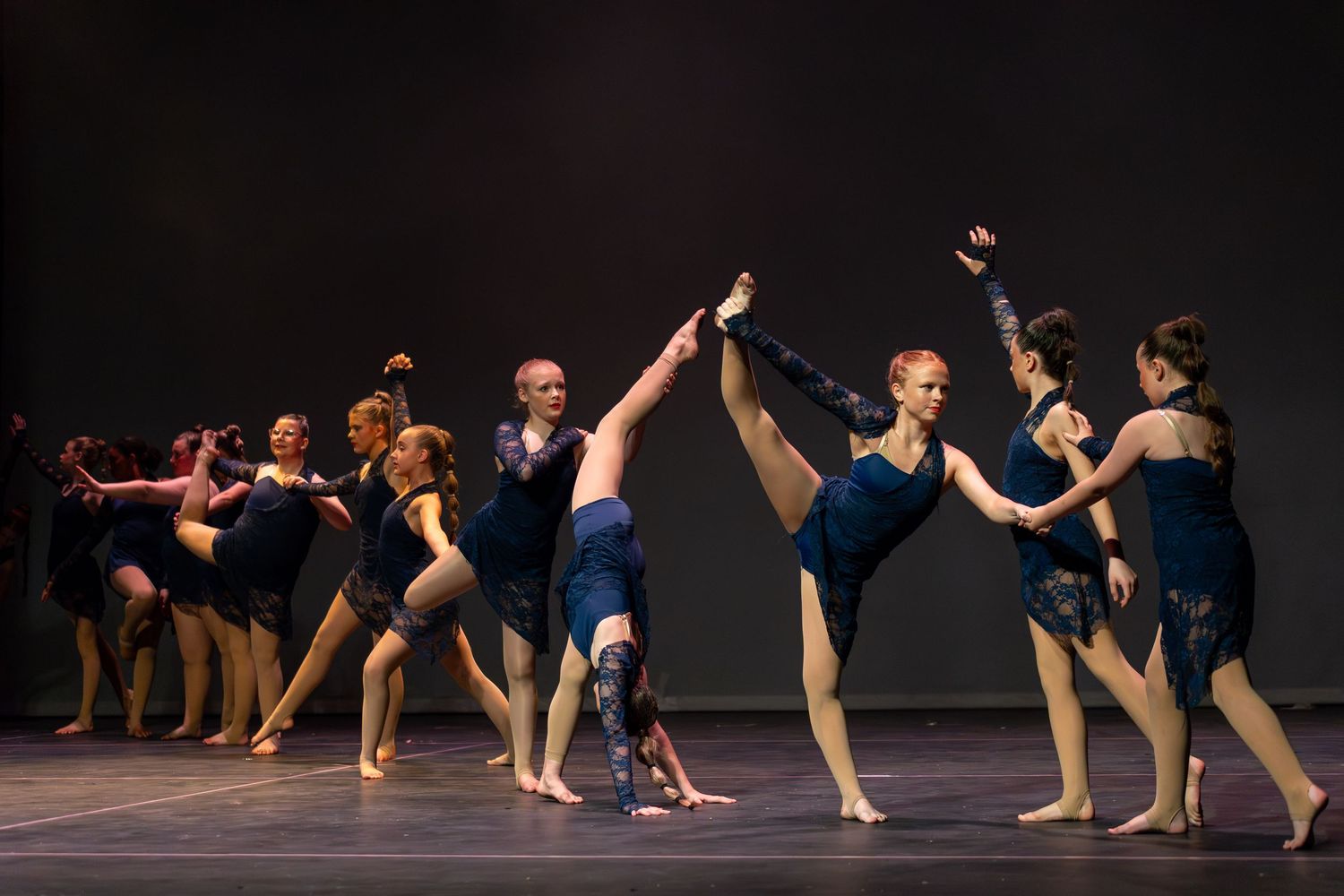 Contemporary dance performers in blue costumes execute elegant poses on a dark stage during a group ballet performance.