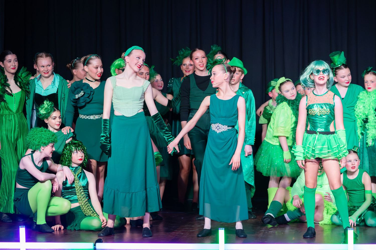 Children and teens perform on stage in coordinated green costumes during a colorful dance recital or theater production.