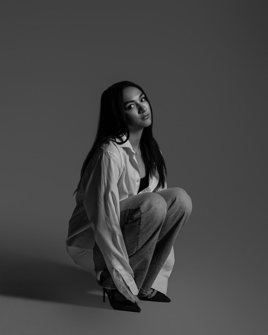 Artistic black and white portrait of person in white shirt and jeans crouching in dramatic lighting.