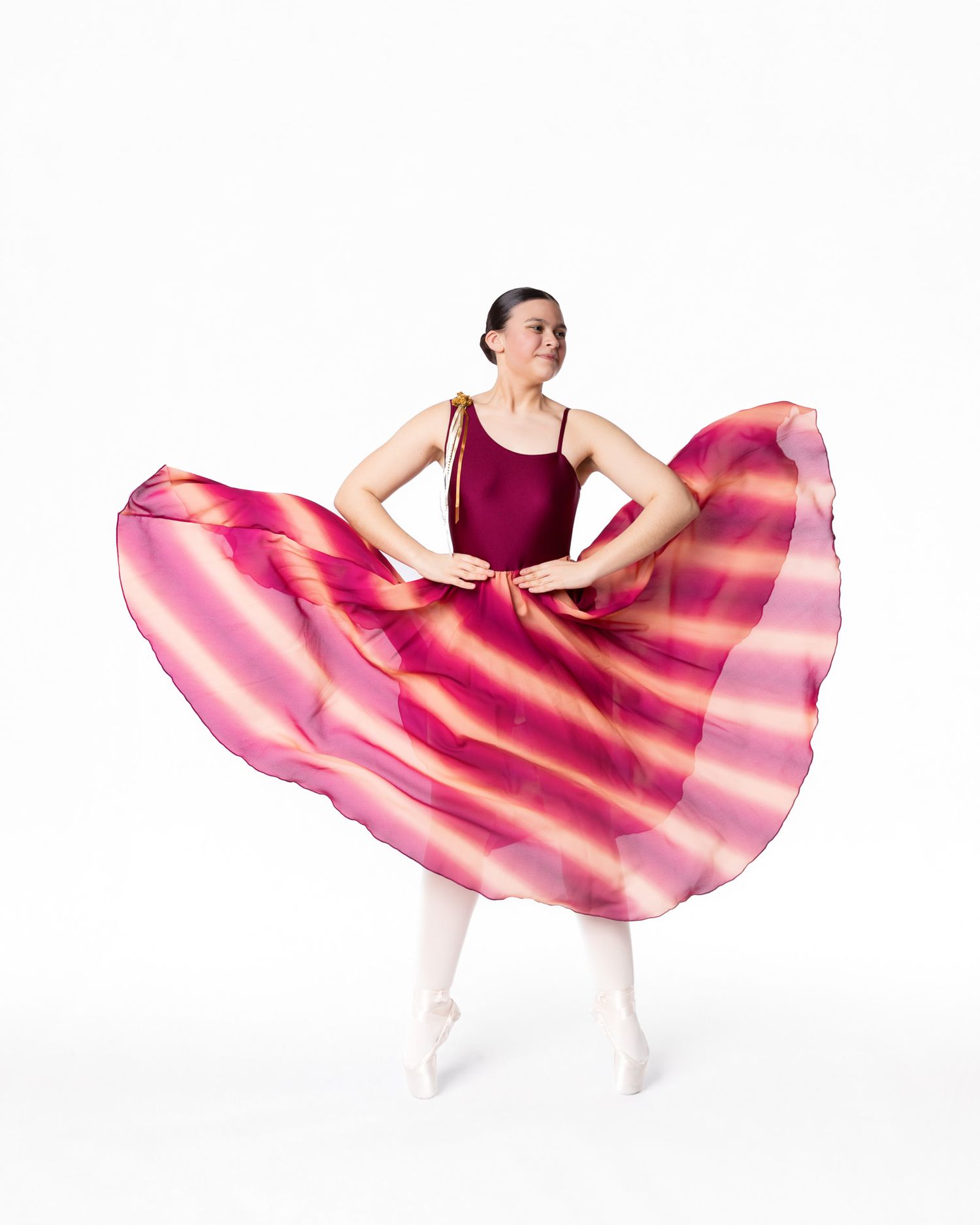 Ballet dancer in burgundy leotard and flowing pink circular skirt poses gracefully in fourth position against white background.