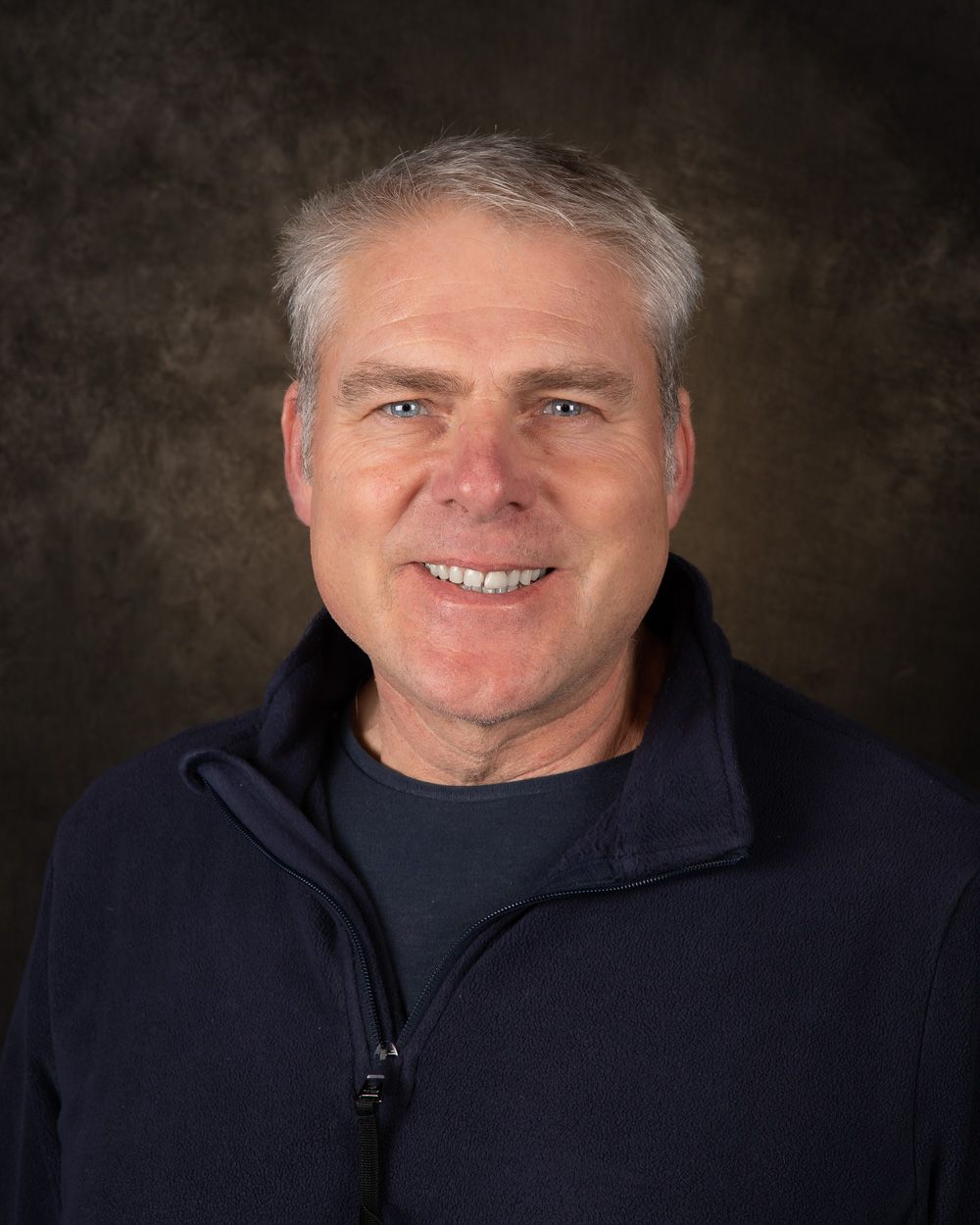 Professional headshot of person in navy jacket against dark background.