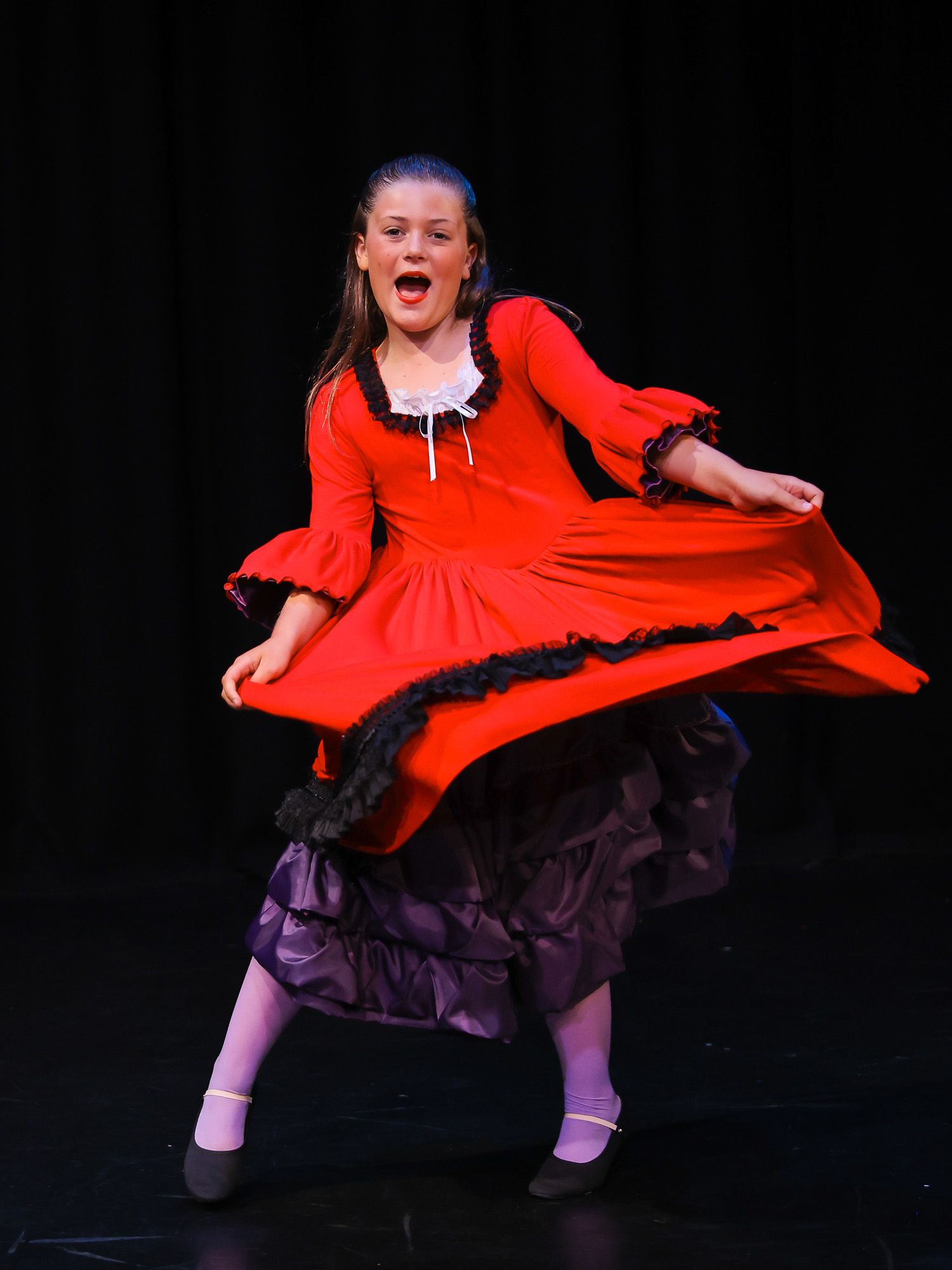 Young girl performing on stage in a red dress with black lace trim, dancing expressively under bright spotlights.