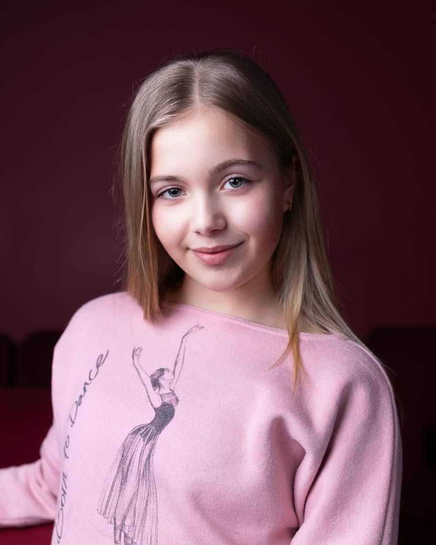 Young girl with blonde hair wearing a pink ballet-themed sweatshirt, posing against a dark red background.