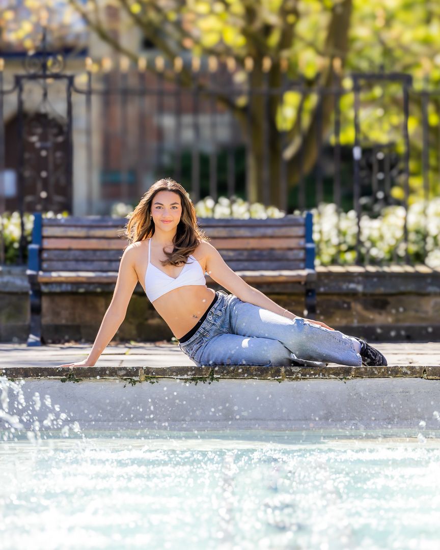 Dancer poses casually by fountain wearing crop top and ripped jeans on sunny day.