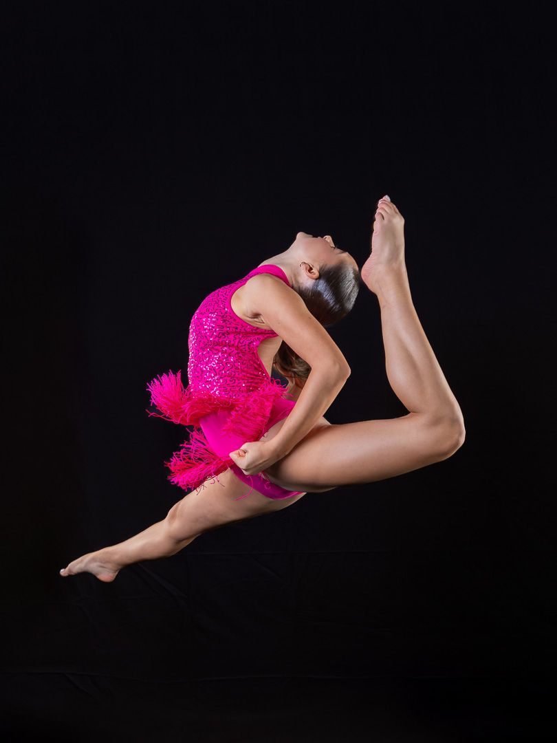 Dancer in bright pink costume performs aerial jump against black backdrop.