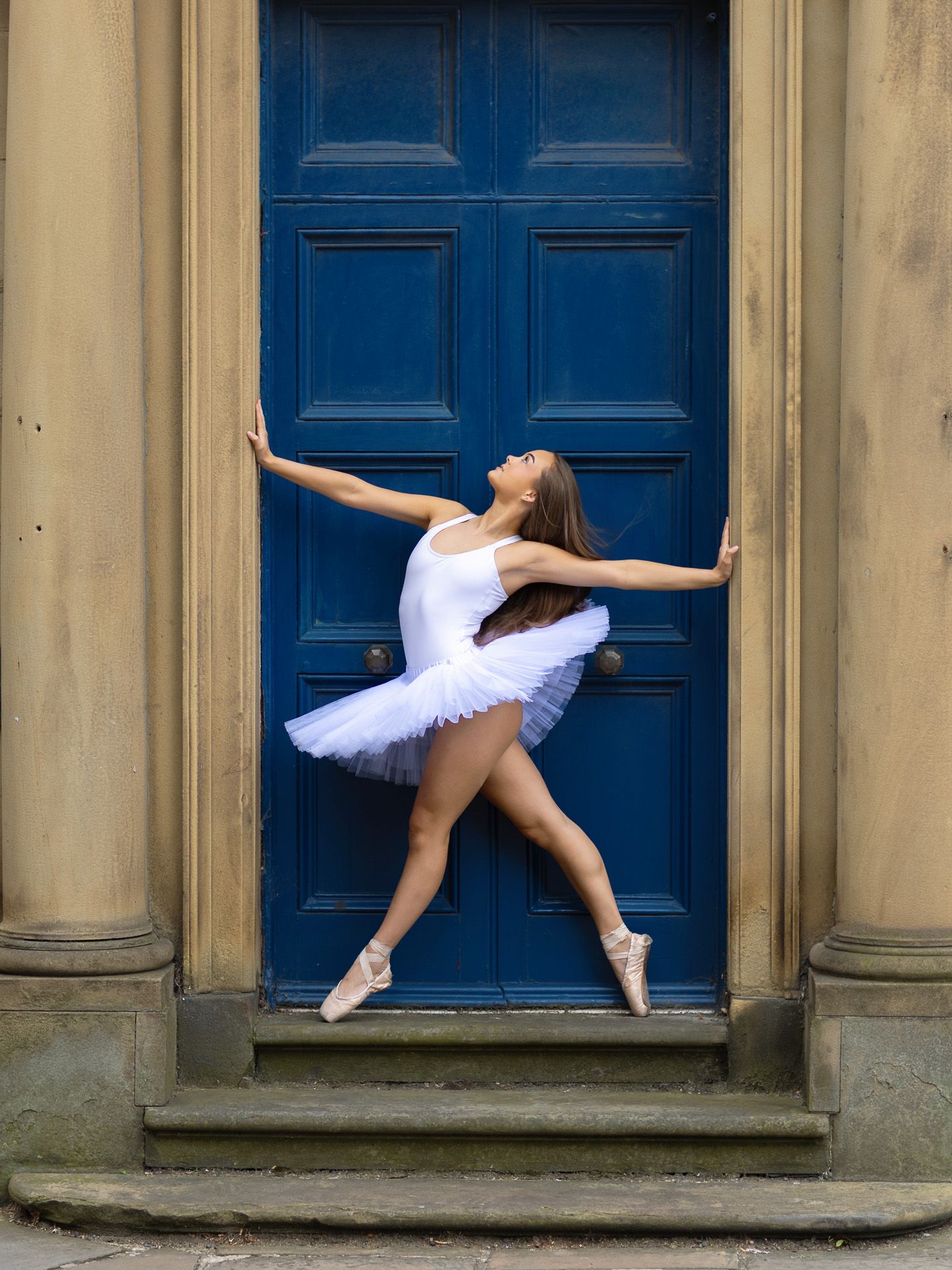 Ballerina in white tutu and pointe shoes posing gracefully in front of a blue door.