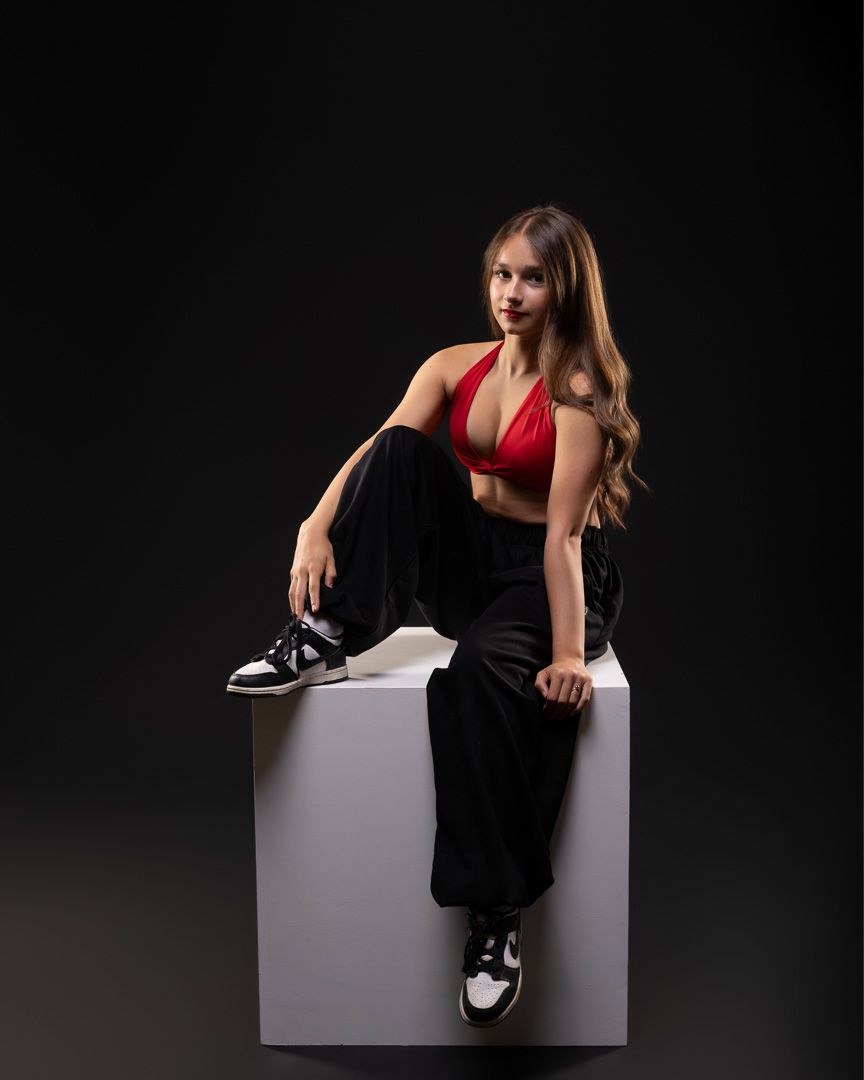 Young woman in red crop top and black pants sits on white cube against dark background wearing black and white sneakers.