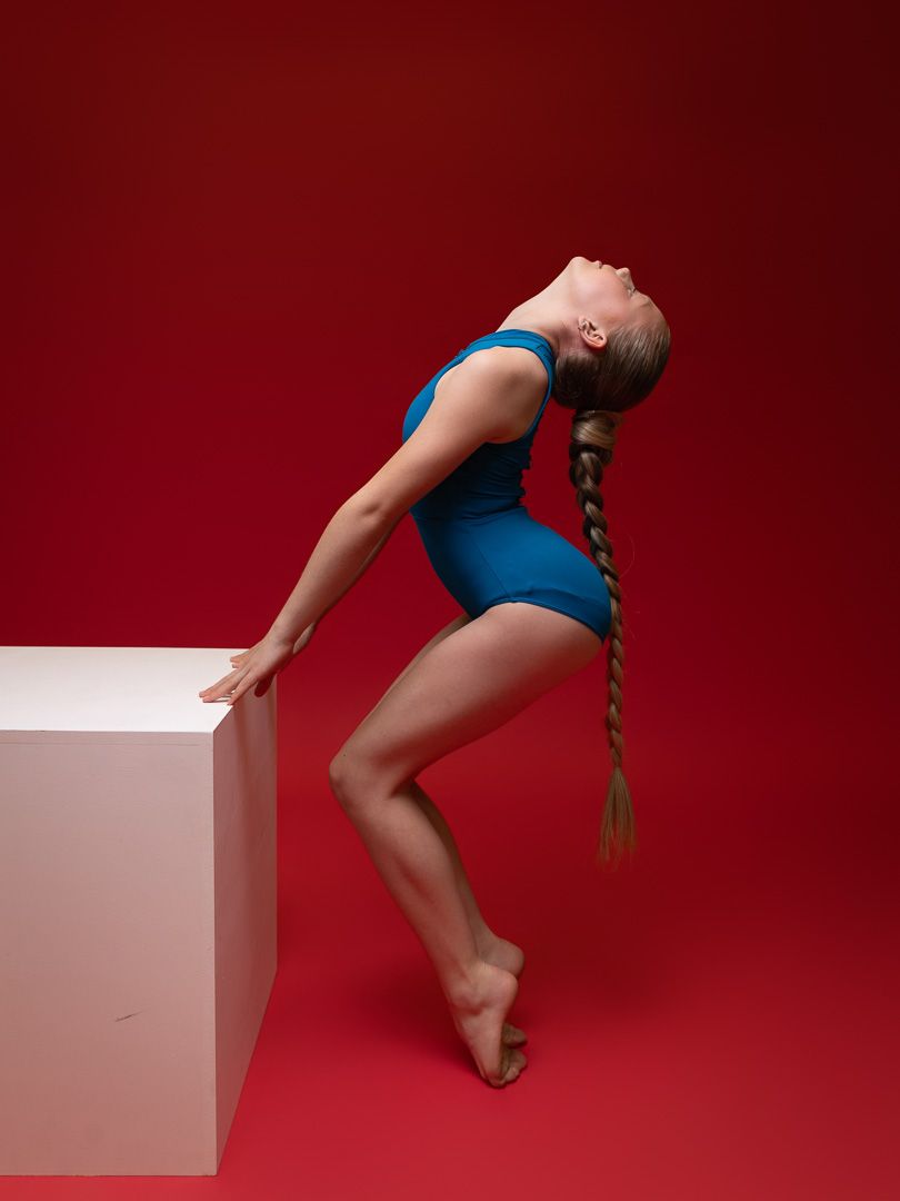 A dancer in a blue leotard with a long braid arches back gracefully against a white cube on a red background.
