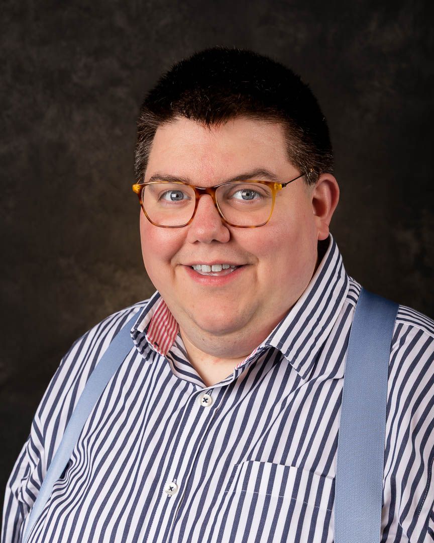 Man smiling in striped shirt with suspenders and yellow glasses, professional headshot photo.