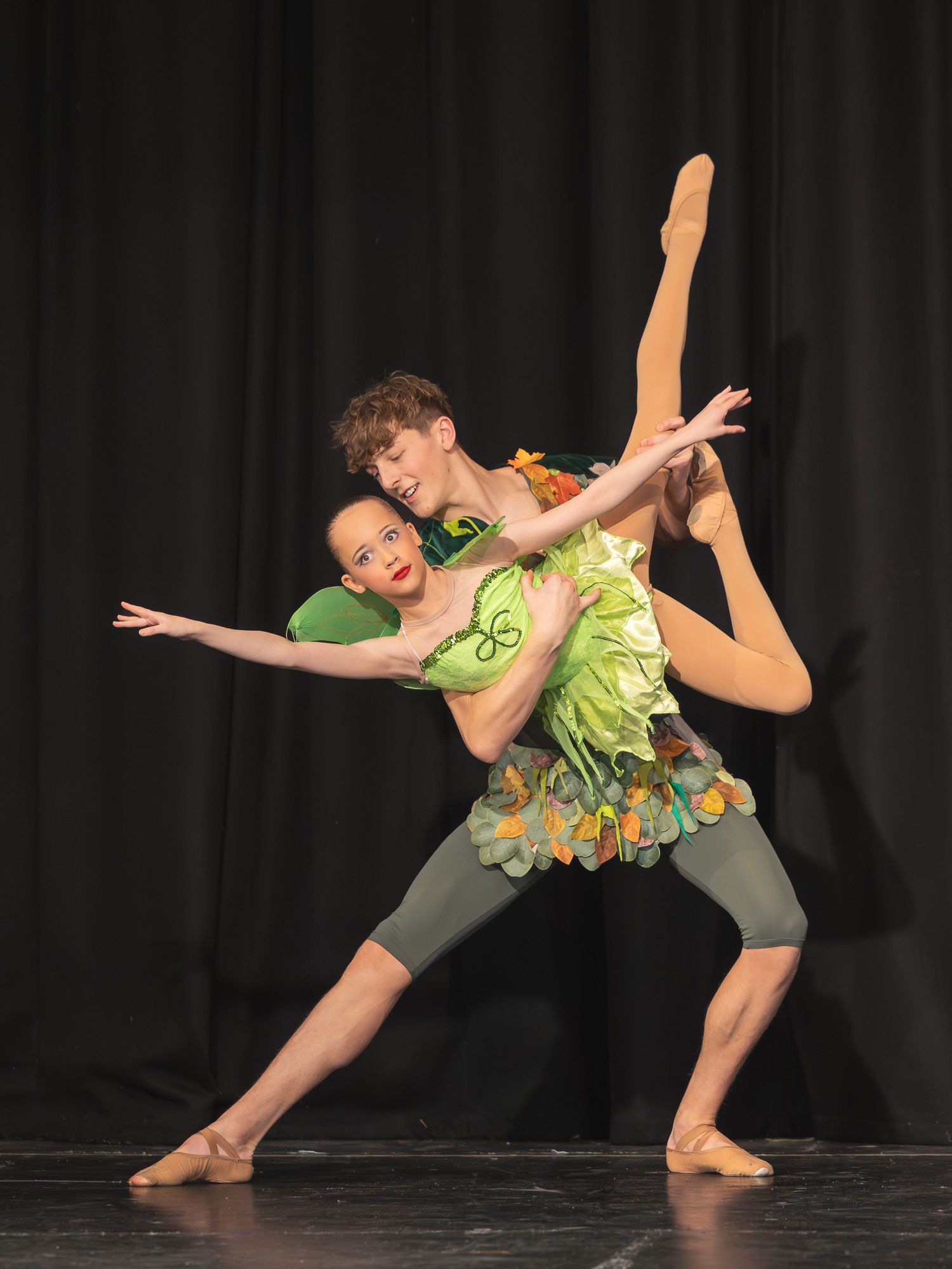 Two dancers in green costumes perform synchronized duet on dark stage.