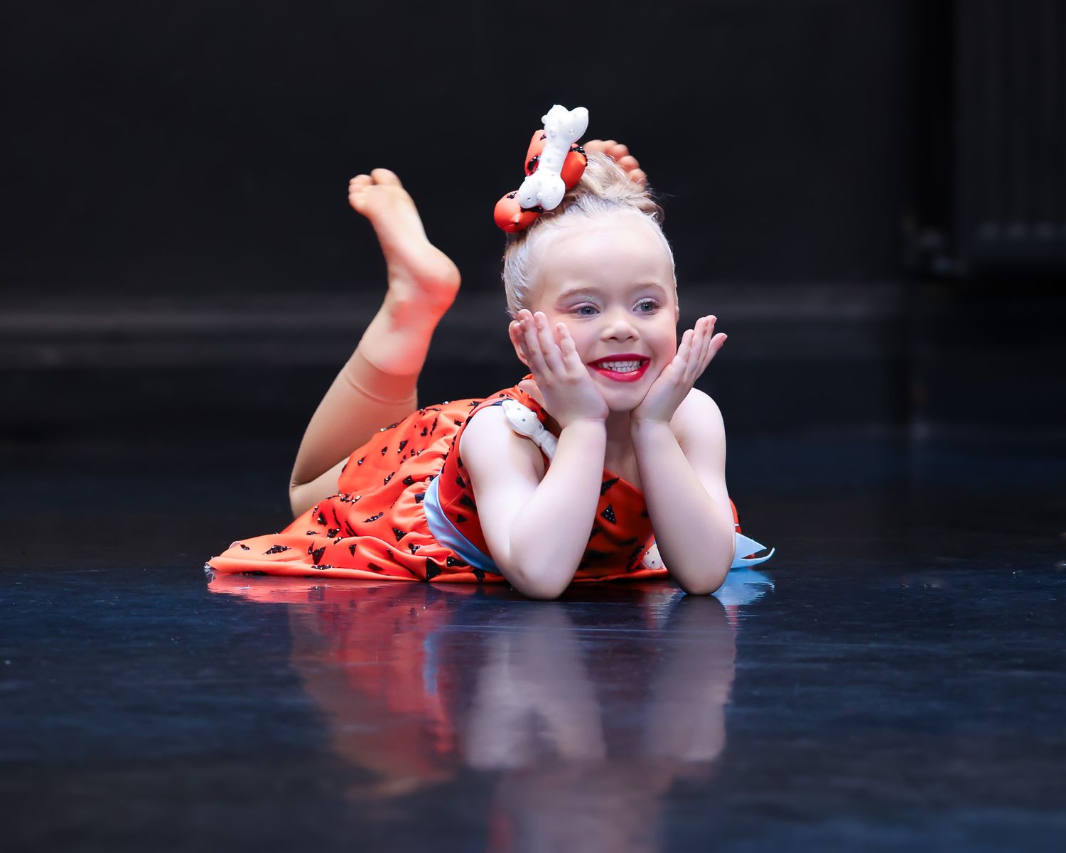 Young girl in red floral dress lying on stage floor, smiling with hands on cheeks, wearing a red bow in her hair.