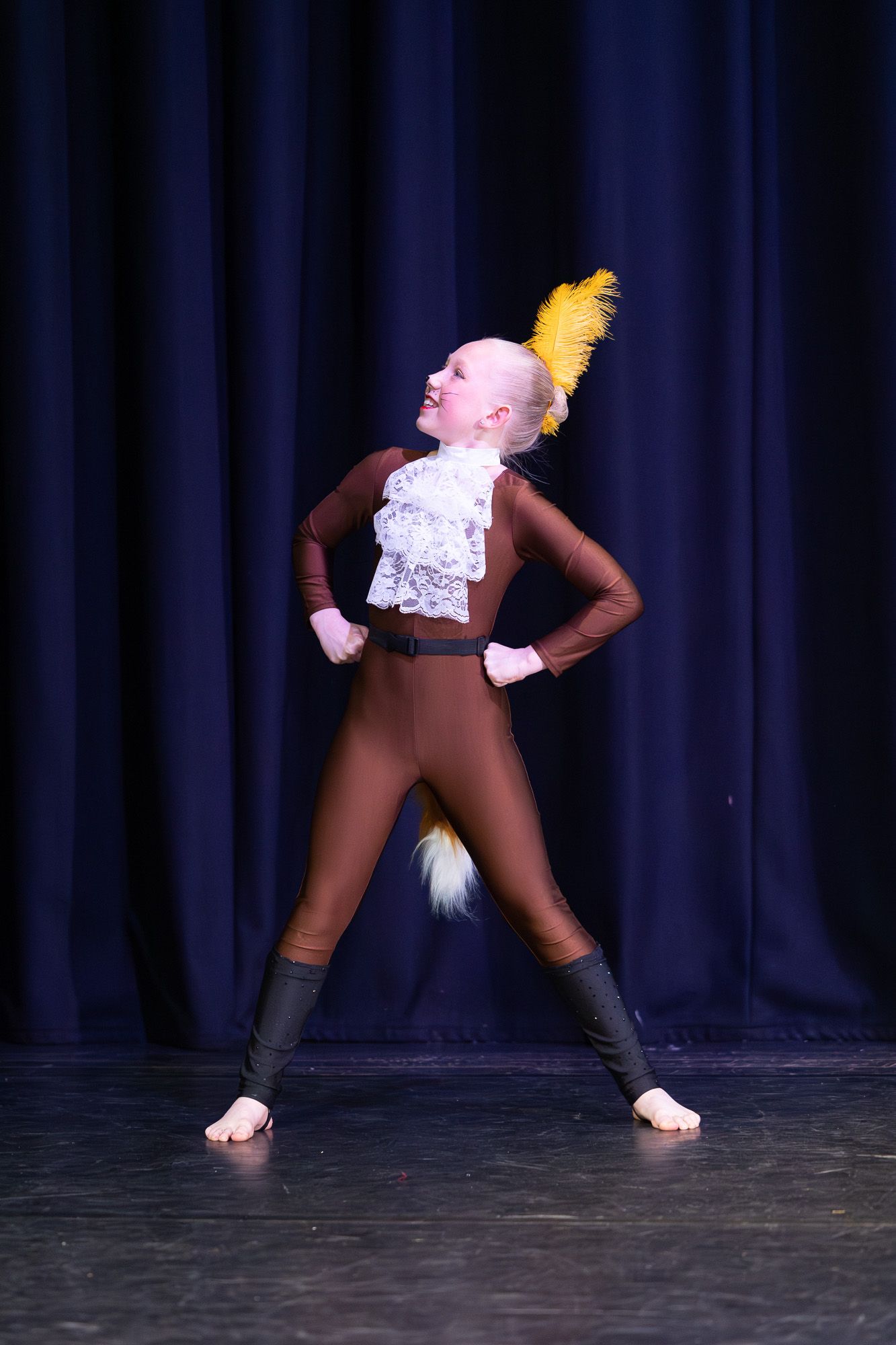 Young dancer in brown costume with yellow feather headpiece and lace collar performs on stage.