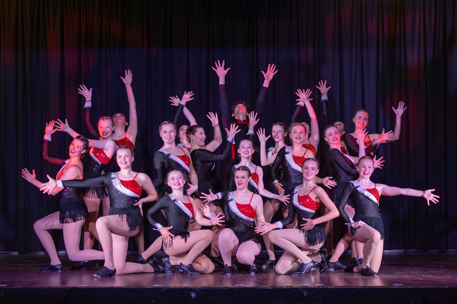 A group of dancers in black and red costumes strike a dramatic pose on stage during a theatrical dance performance.