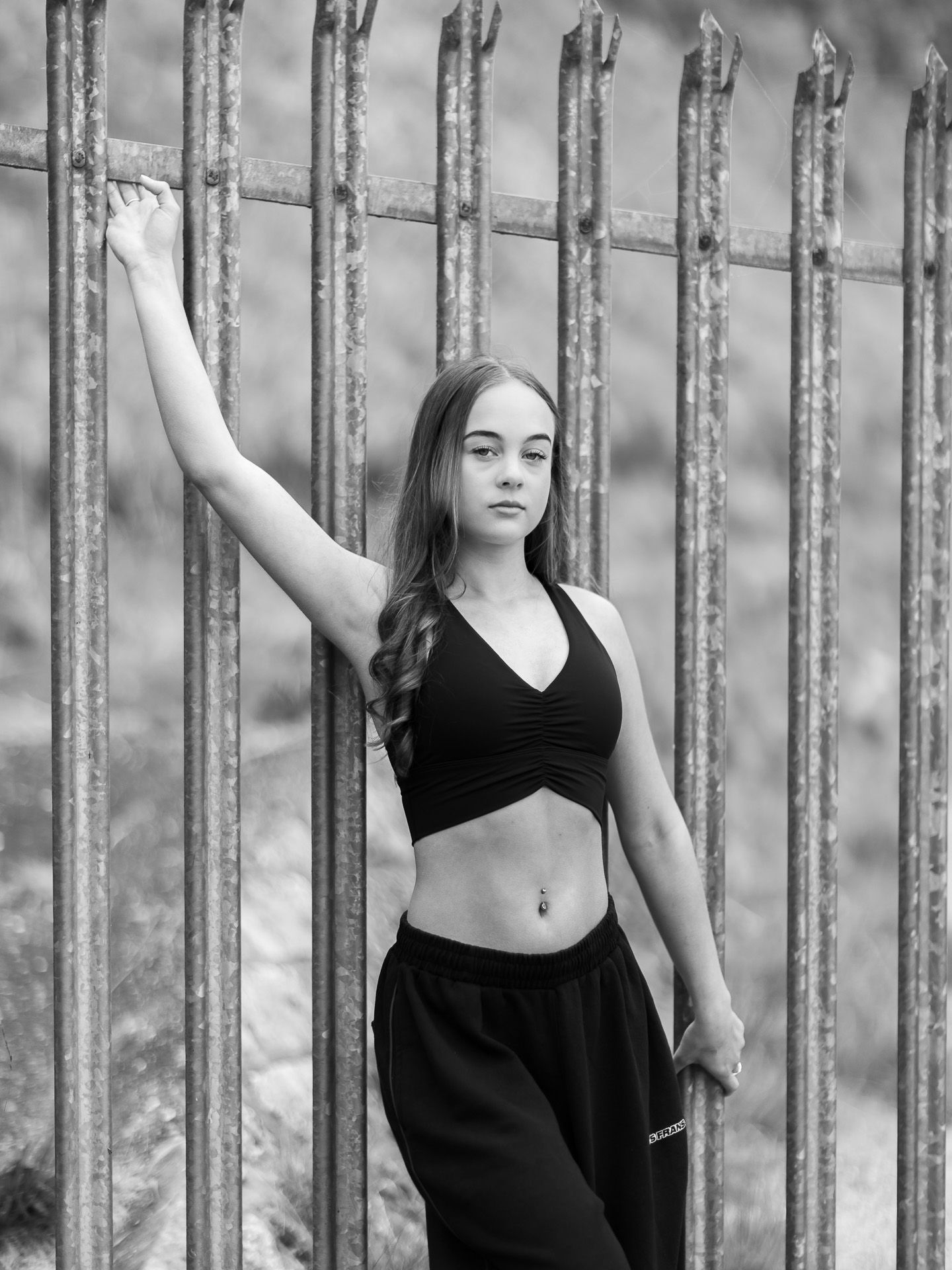Black and white photo of a young woman in a crop top posing against a metal fence outdoors.