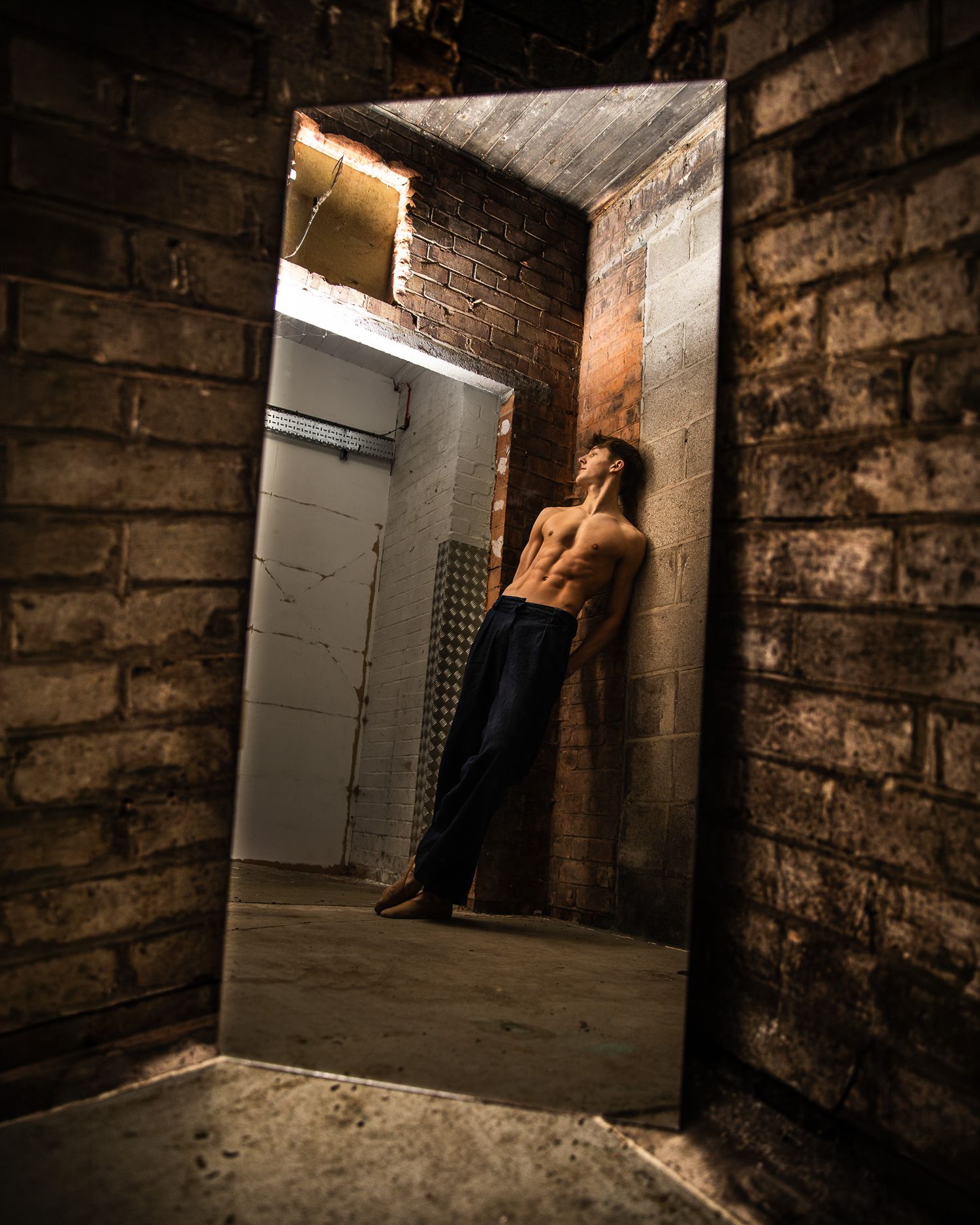Shirtless athletic man leaning against brick wall in dramatic doorway, illuminated by warm light in an urban setting.