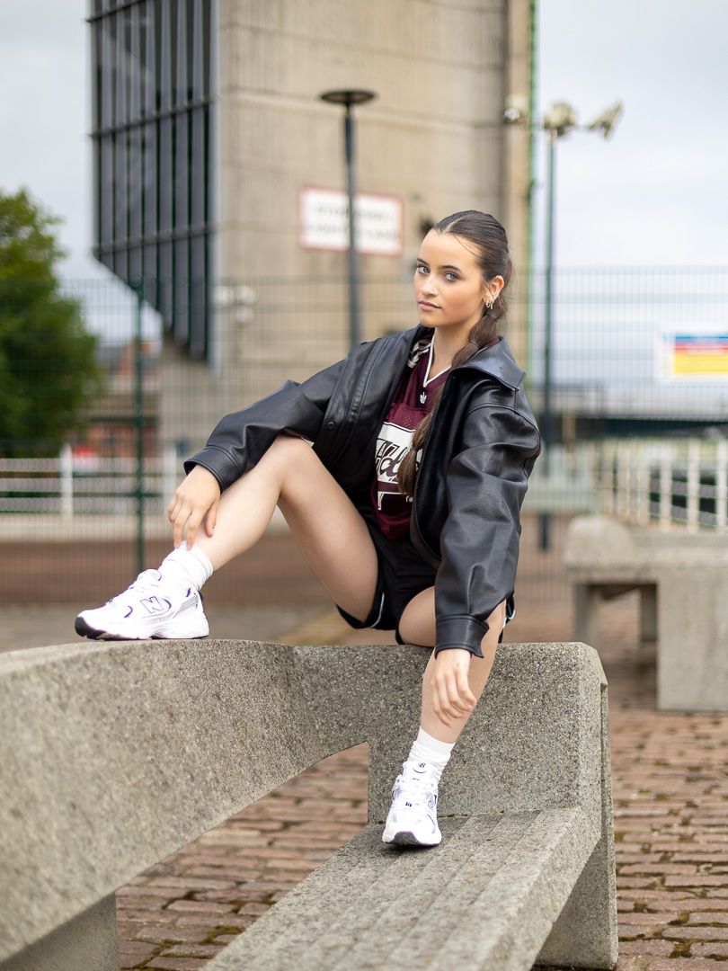 Young woman in leather jacket, mini skirt, and white sneakers sitting on concrete bench in urban setting.