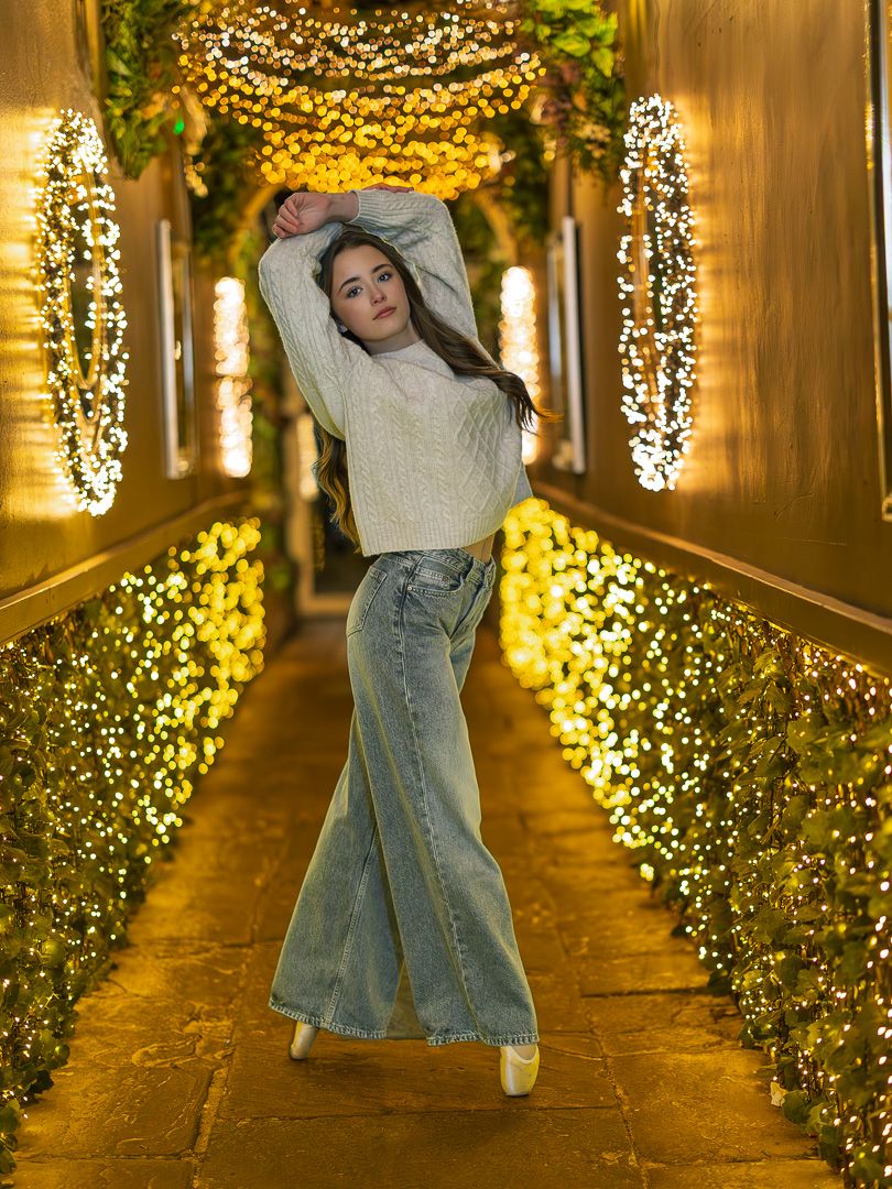 A person in a cream sweater and jeans poses in a festive hallway decorated with twinkling string lights and greenery.