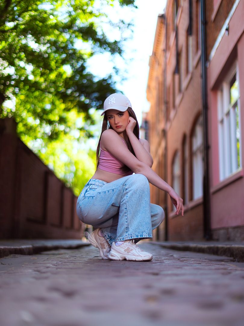Stylish young woman in wide-leg jeans and white cap crouching on cobblestone alley between brick buildings.