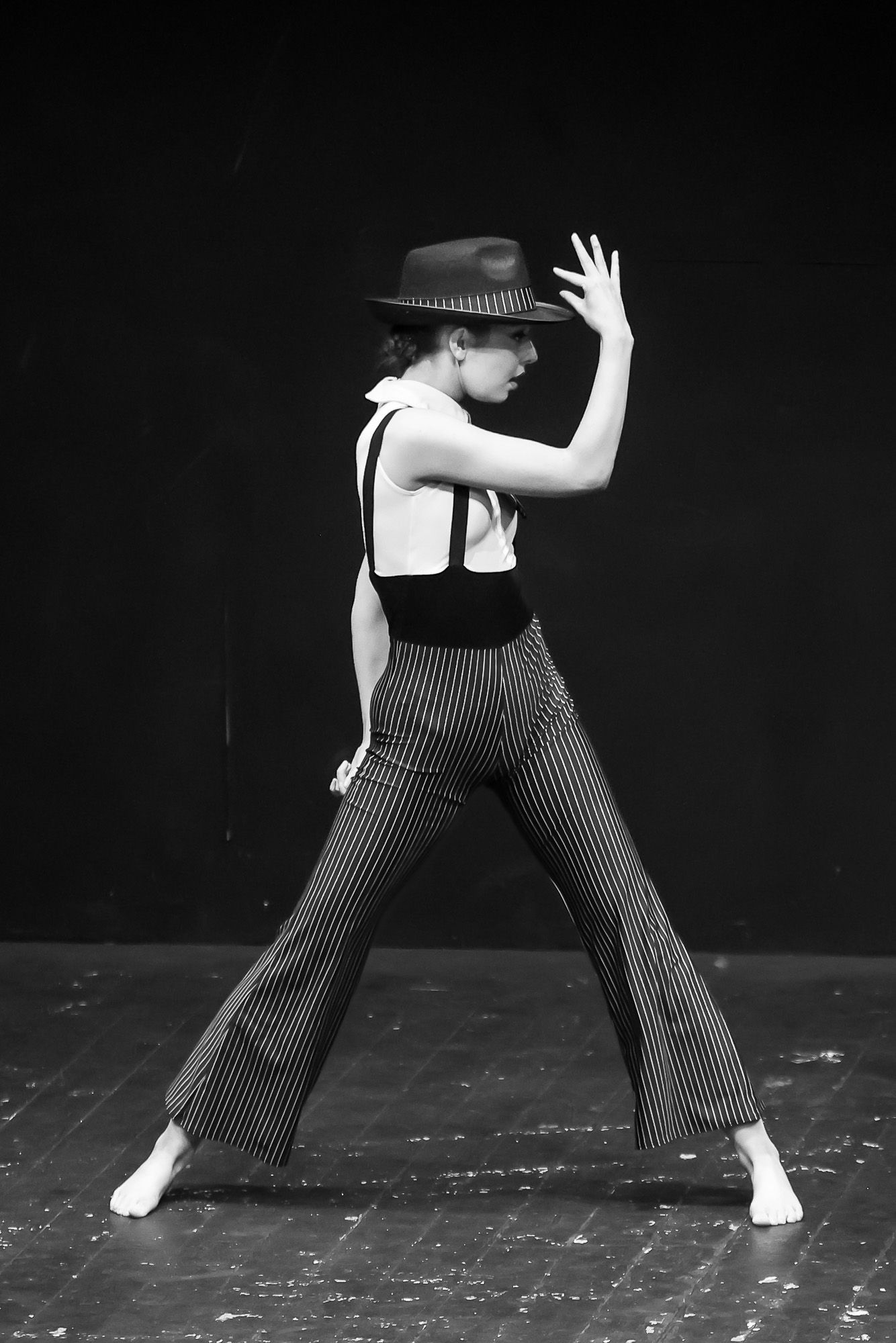 Black and white photo of a dancer in striped pants, suspenders, and fedora hat performing an elegant pose on stage.