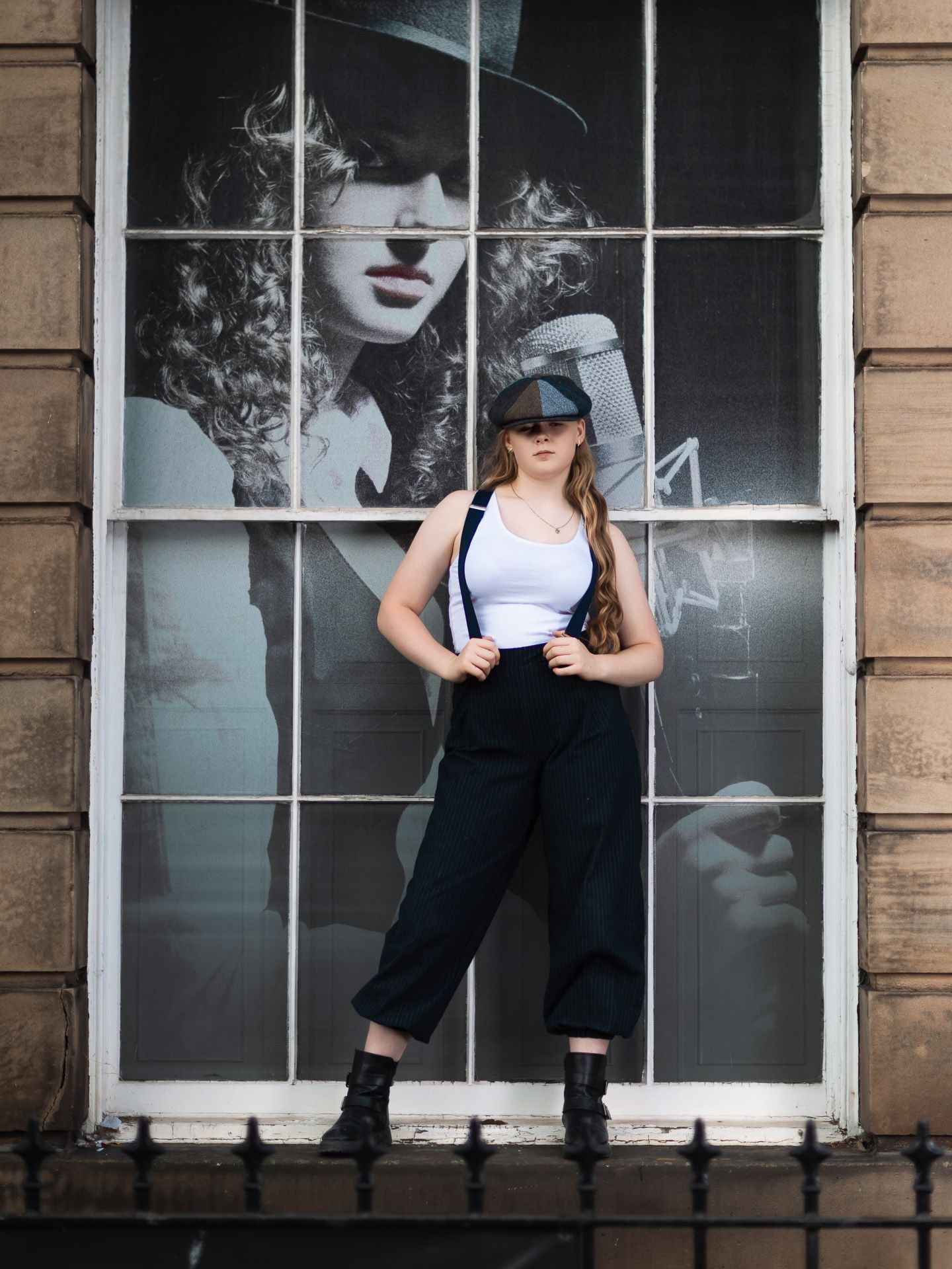Young woman in white top, black suspenders, cap, and boots poses before a large window with monochrome portrait.