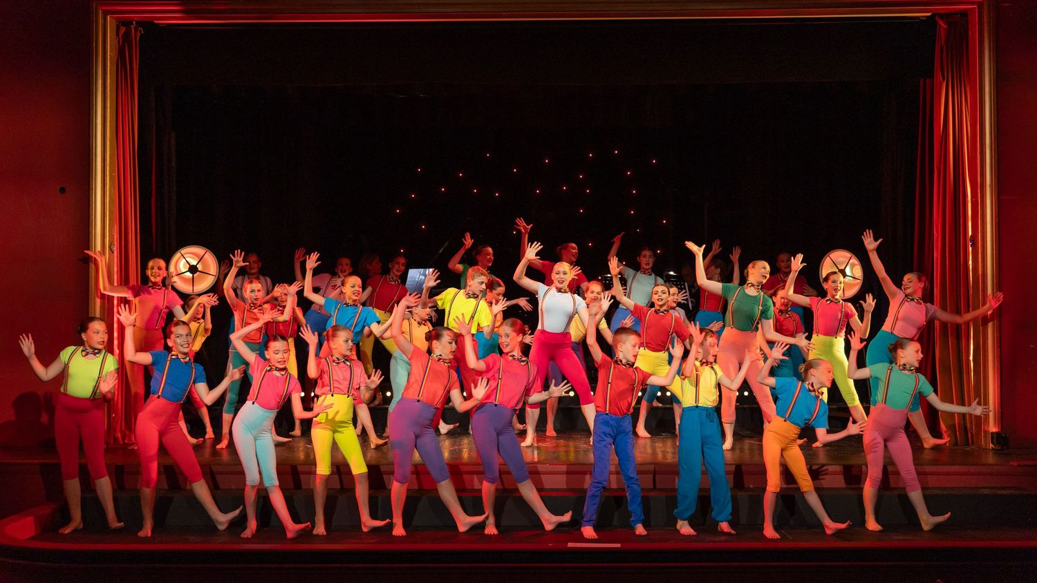 Colorful theatrical dance performance with large ensemble cast in vibrant costumes on a stage with red curtain backdrop.