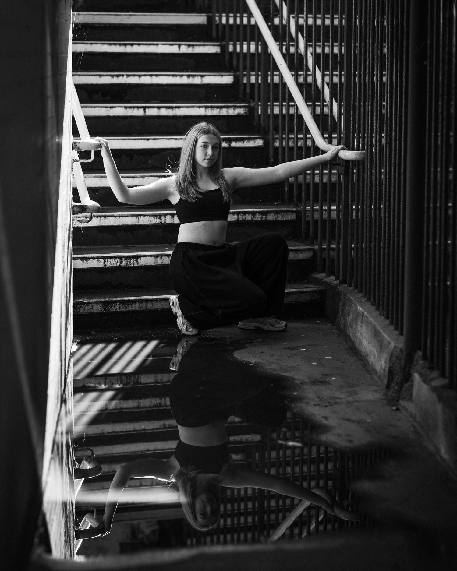 Woman in black outfit poses gracefully on urban metal stairs in dramatic black and white photography.